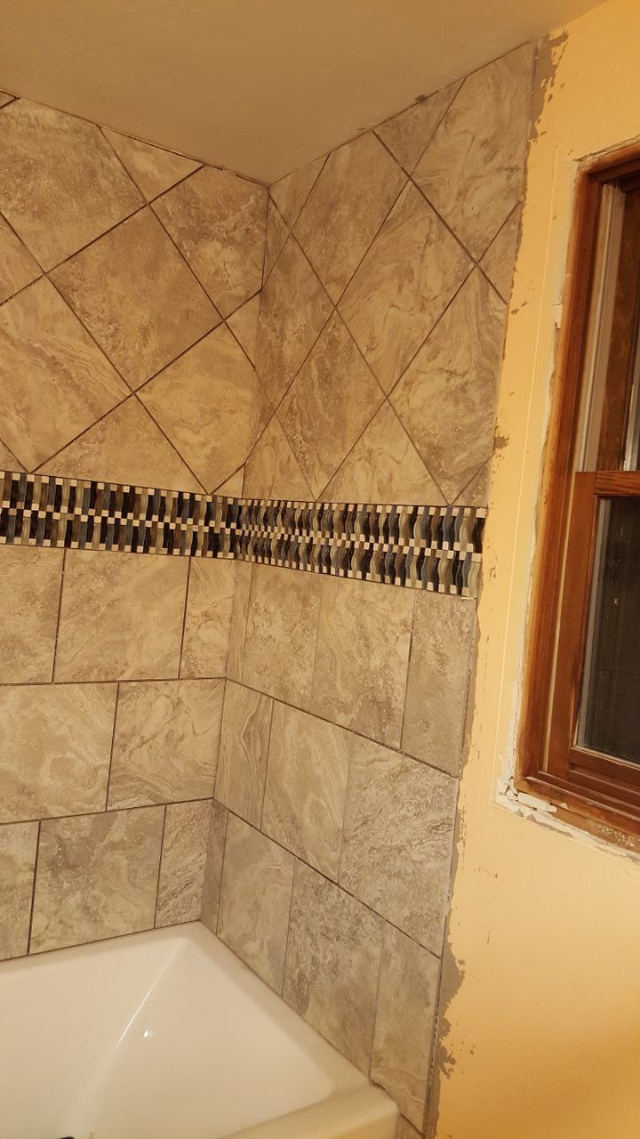 Tiled shower corner with light brown tiles in a diamond pattern. A dark band of mosaic tiles accents the design near the middle, next to a window.
