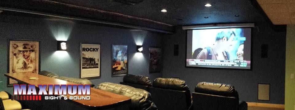 Home theater with movie posters, a bar, and a screen playing a film. Blue wall, black seating, and ceiling lights.