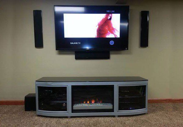 TV mounted on a beige wall with speakers and a media console featuring a fireplace and sound system.