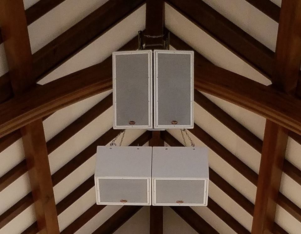 Four white rectangular speakers mounted on a wooden beam ceiling.
