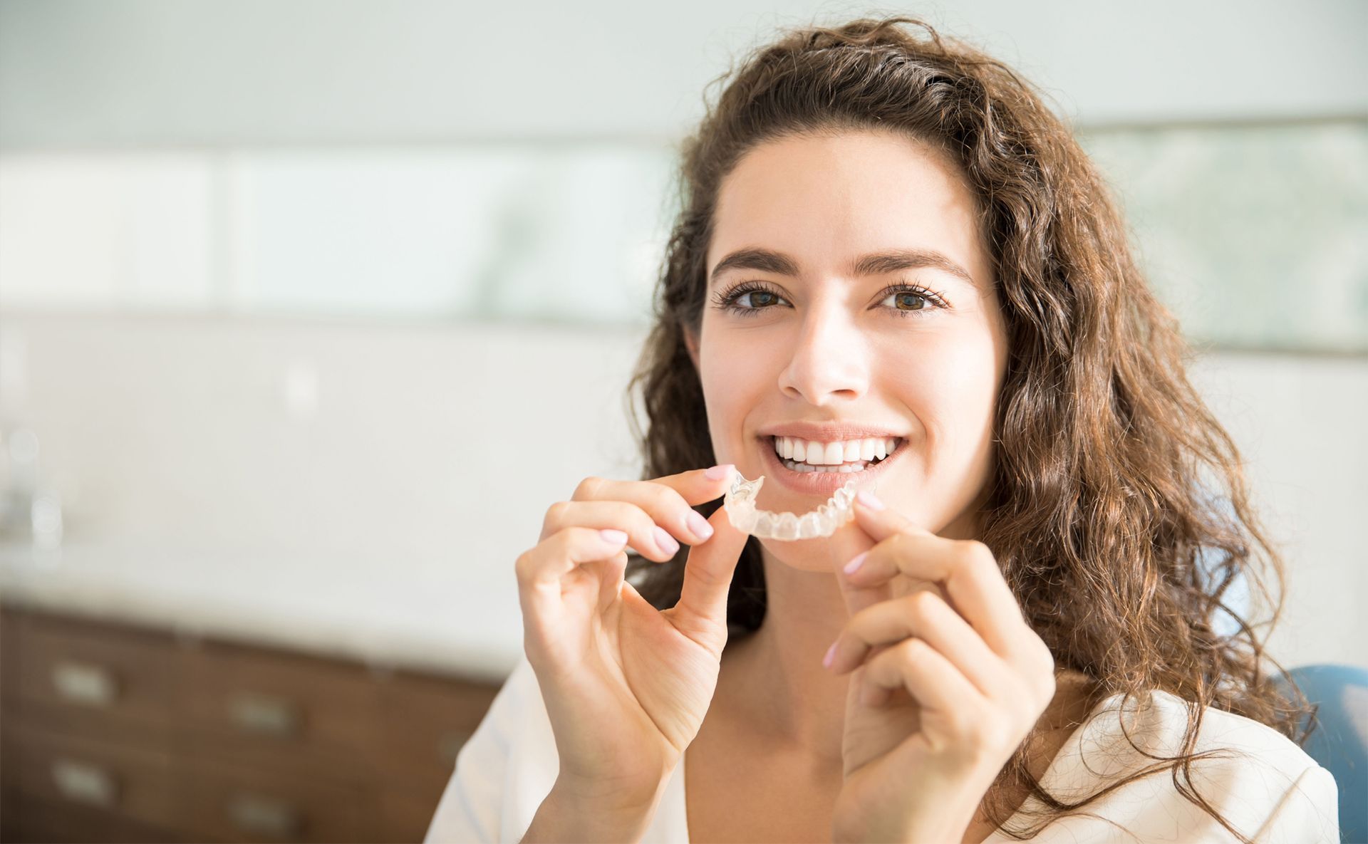 Woman holding clear teeth aligner, smiling, in a dental office setting.