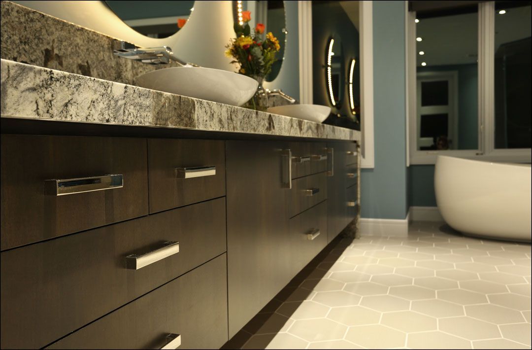 Bathroom vanity with dark brown cabinets, stone countertop, and white sinks. A white tub is in the background.