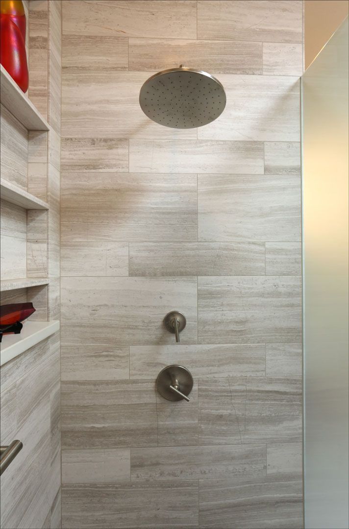 Shower with light-colored wood-look tiles, a round shower head, and brushed metal fixtures. A shelf is visible on the left.
