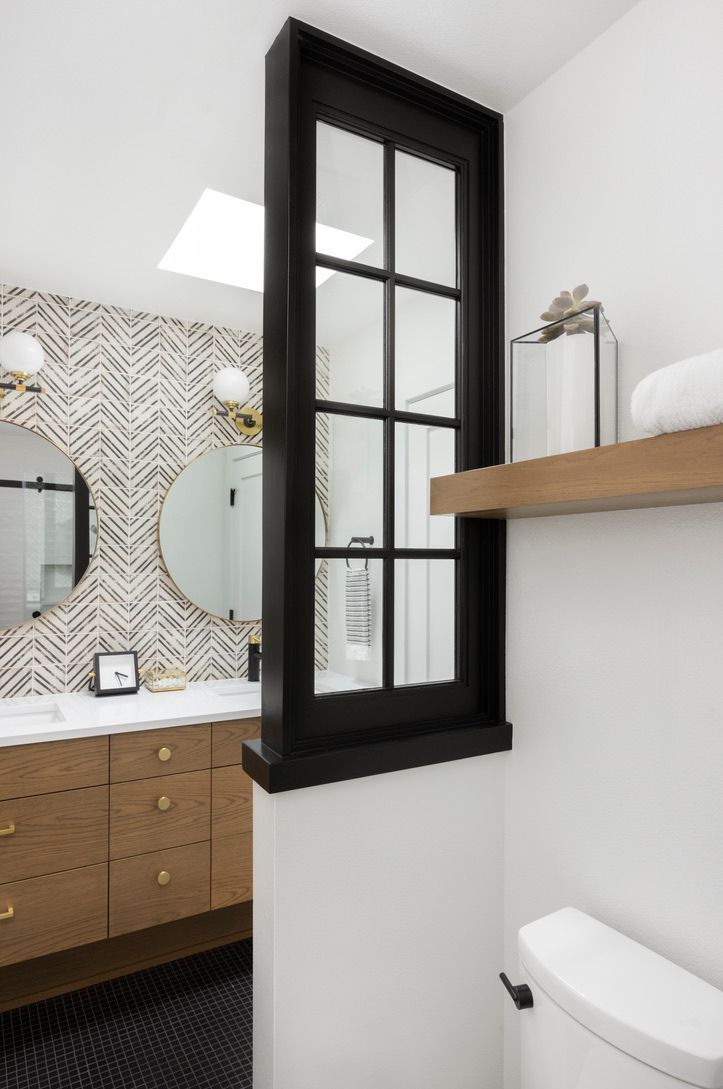 Bathroom with a black-framed window separating the toilet and vanity area; white walls and wooden shelf.
