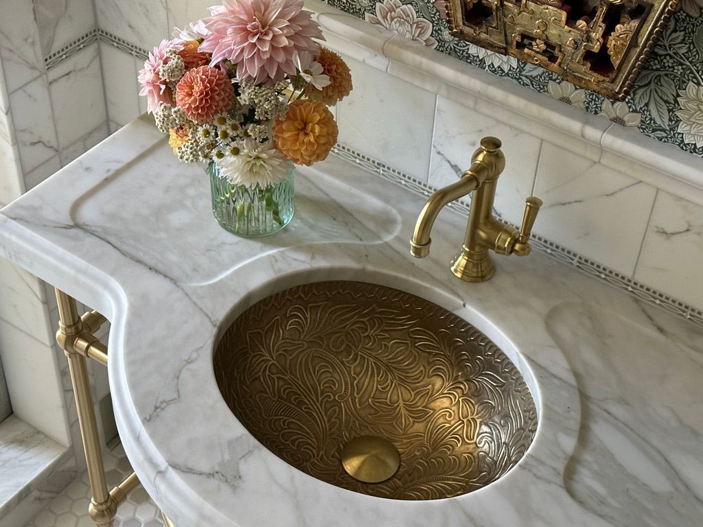 Marble vanity with brass sink, faucet, and vase of flowers.