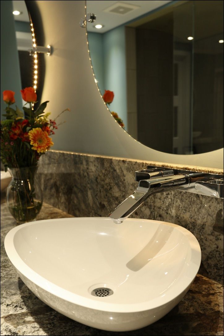 White oval sink with running water in a modern bathroom. Dark counter, large mirror, vase of flowers.