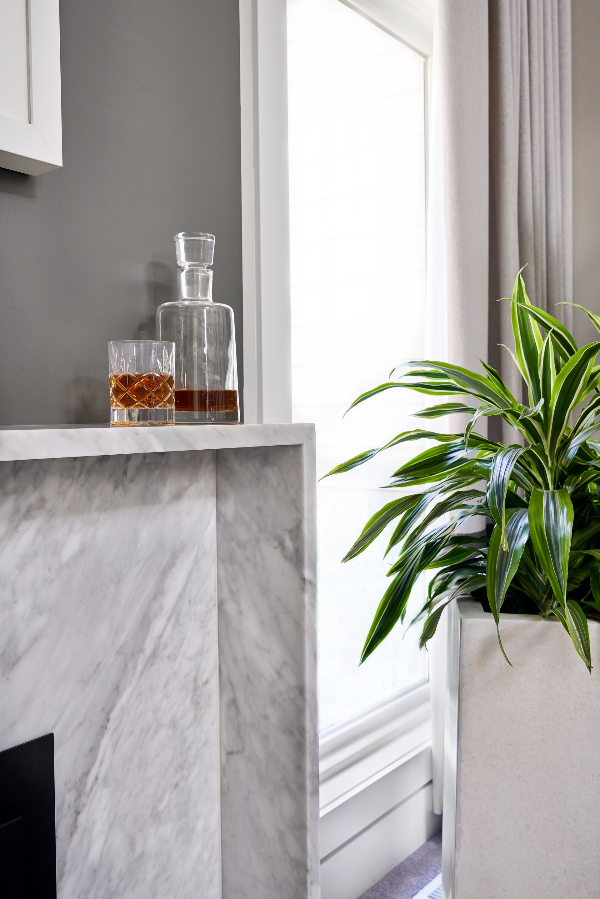Whiskey decanter and glass sit on marble shelf near a window, with a potted plant nearby.