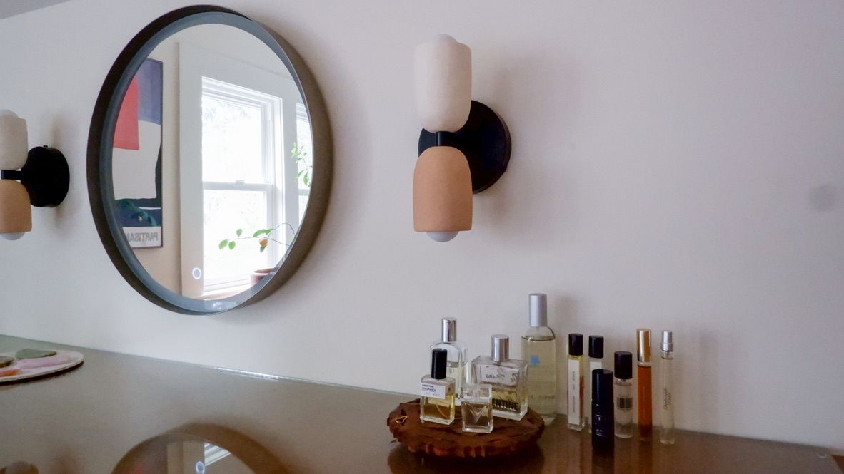 A vanity with a round mirror, modern sconces, and perfume bottles on a wooden tray.
