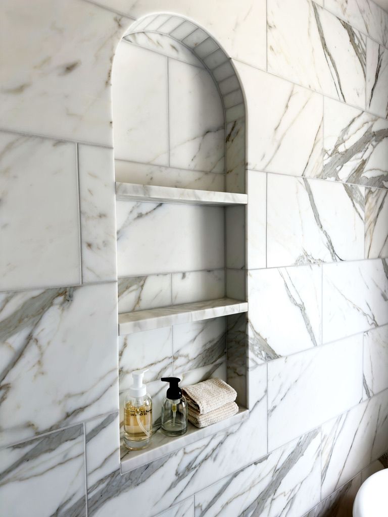 Marble tiled shower niche with arched top, holding toiletries and towels.