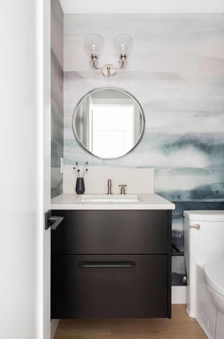 A modern bathroom with black vanity, round mirror, patterned wallpaper, and a white toilet.