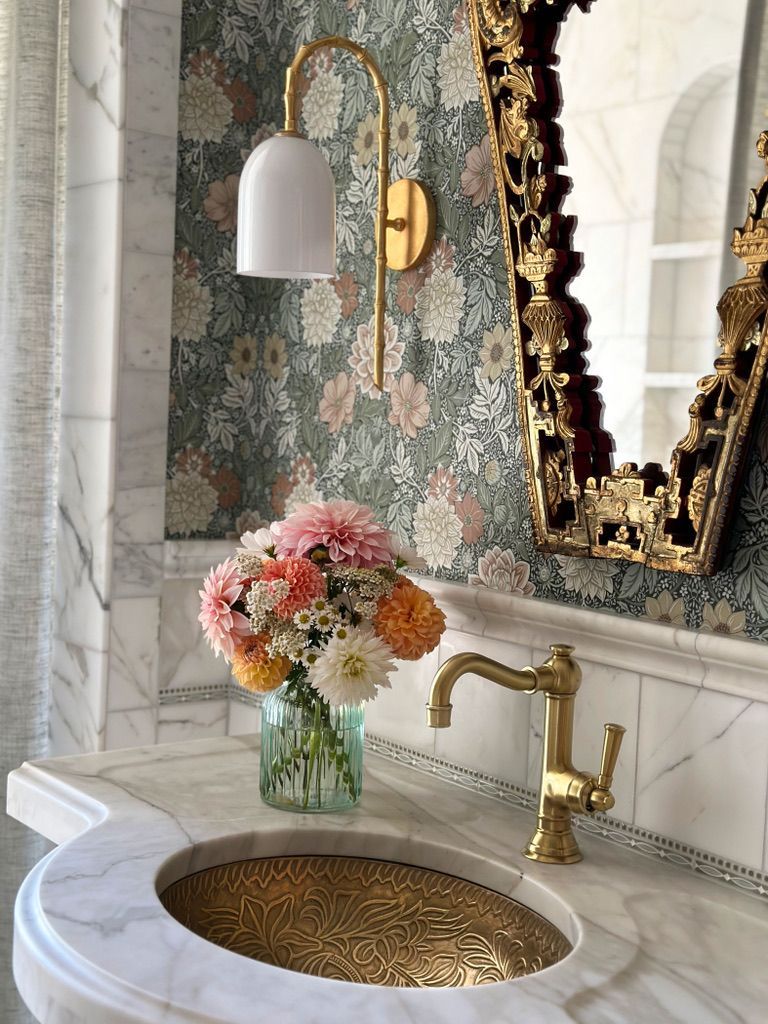 Elegant bathroom with floral wallpaper, marble countertop, gold sink, and flowers.
