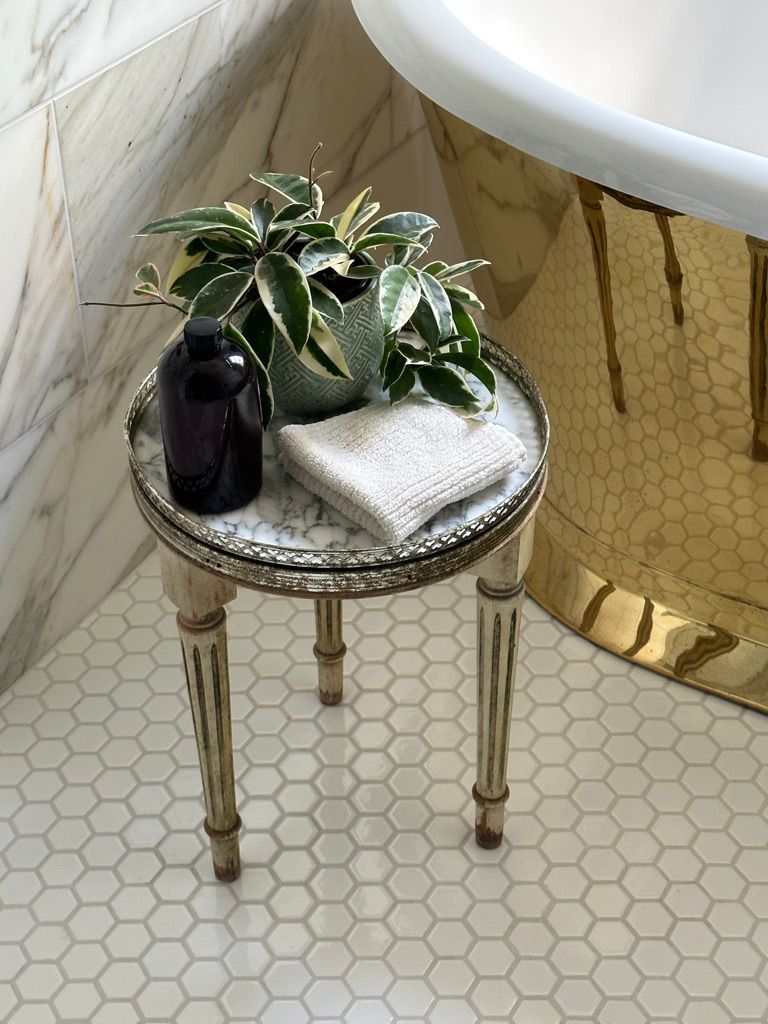 Small table with bath accessories: plant, bottle, washcloth. Near a gold tub and marble tile.