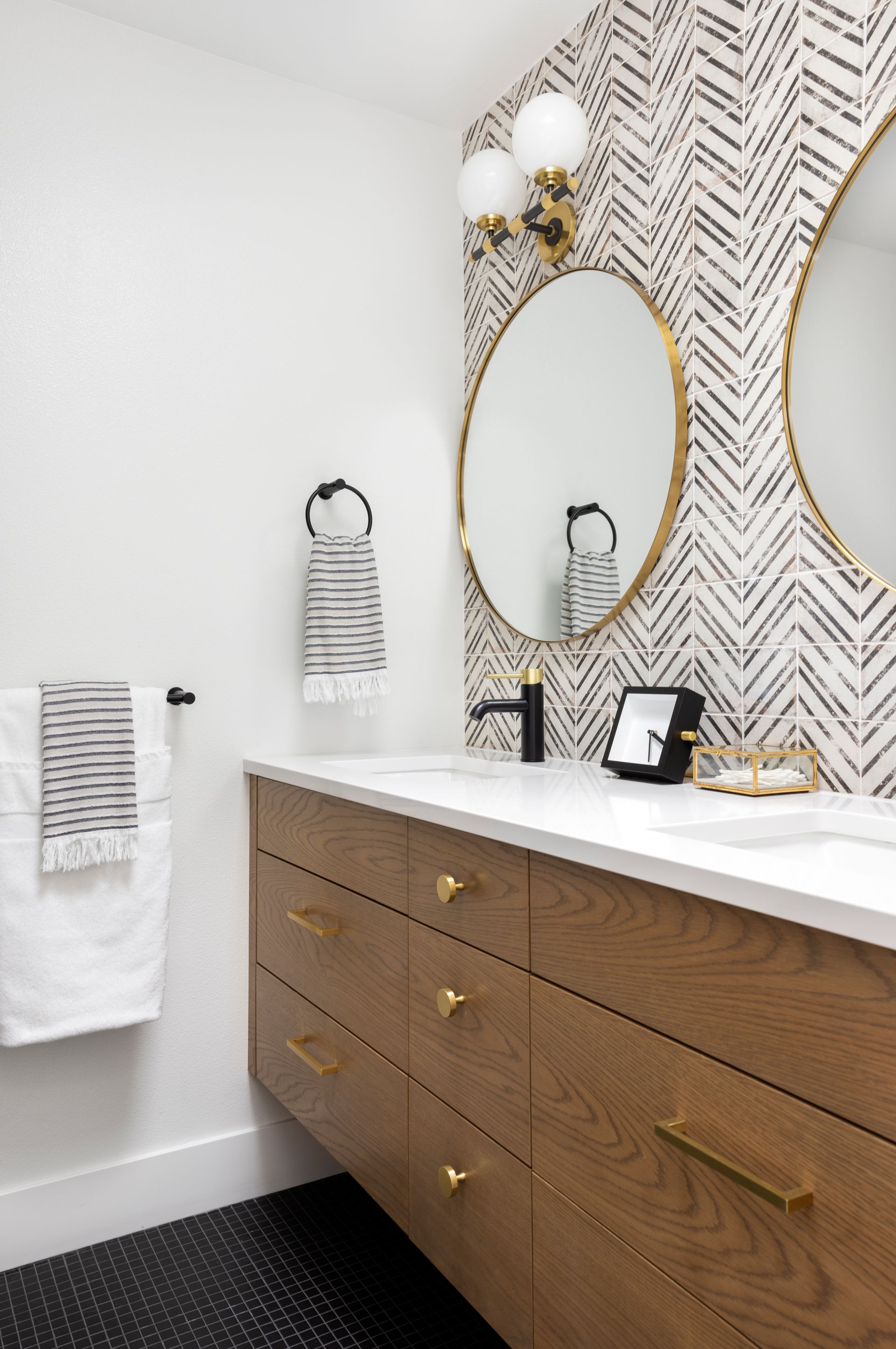 Bathroom with wood vanity, patterned accent wall, round mirrors, and gold fixtures.