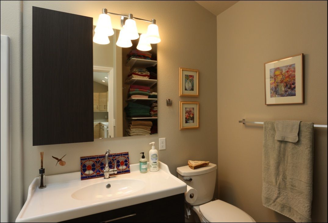 Bathroom with dark vanity, white sink, mirror, toilet, towels, artwork, and shelves of folded towels.