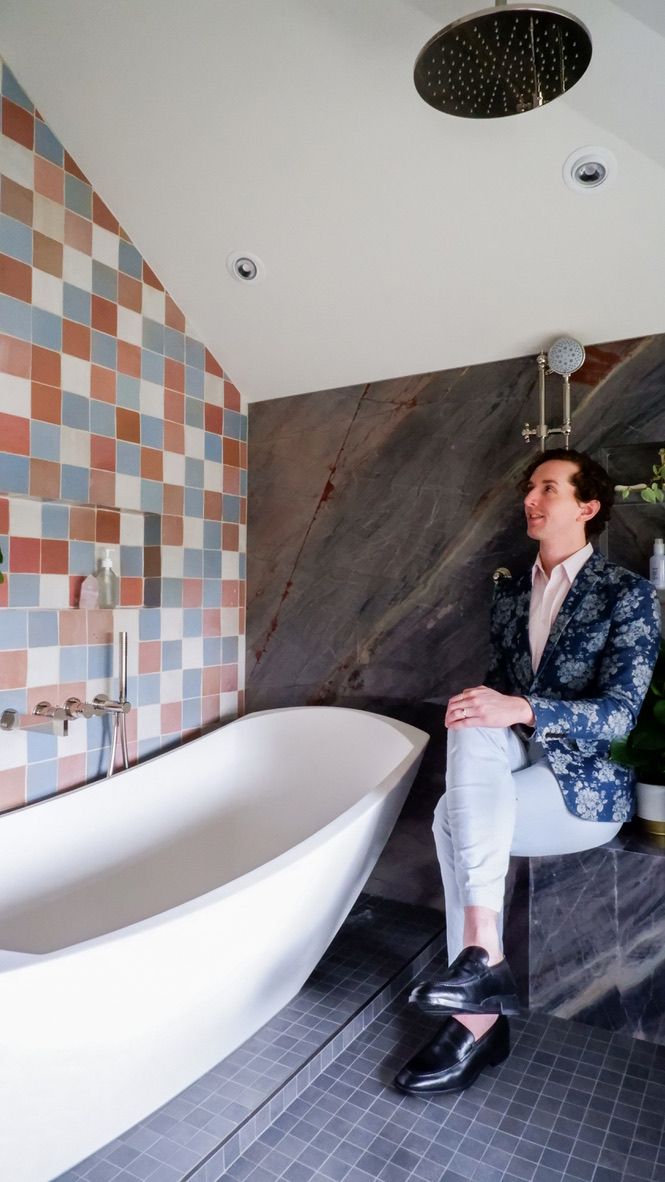 Man in blue jacket sits near a white bathtub in a modern bathroom with tiled walls.