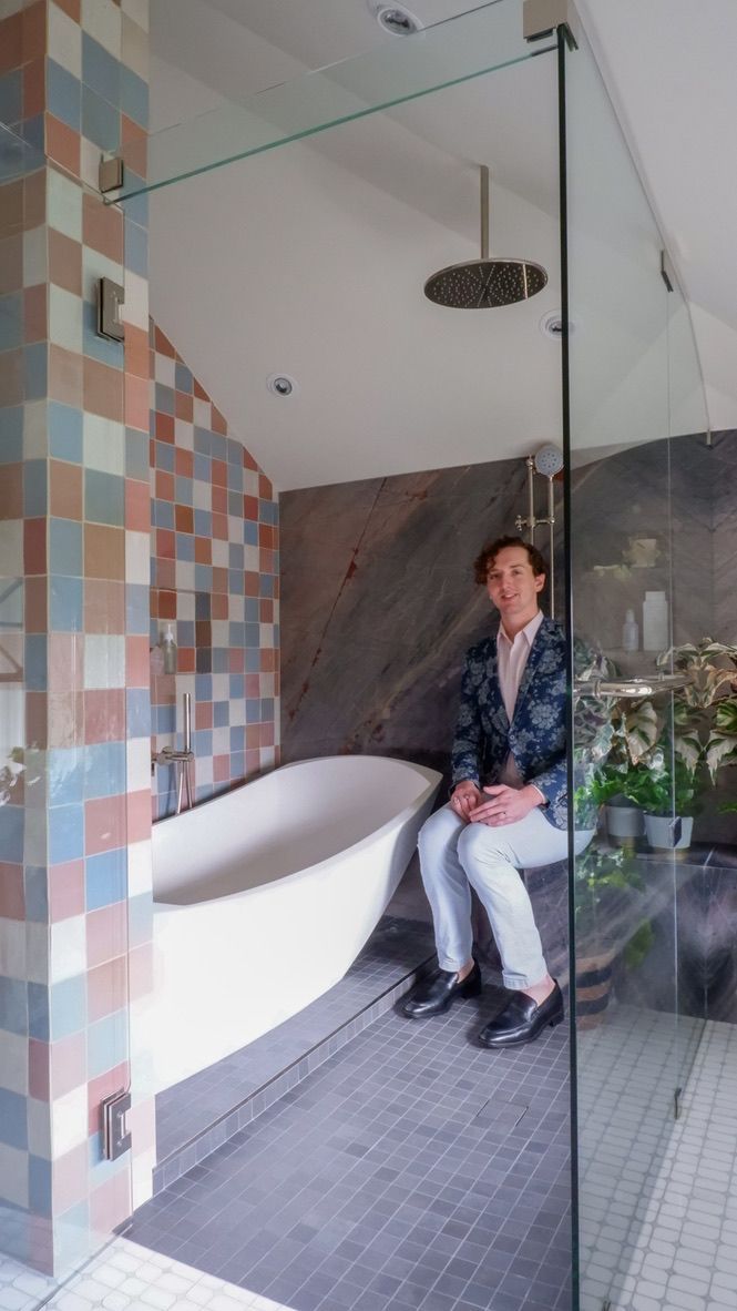 Man sitting in a bathroom with a tub and shower. Multi-colored tile, glass, and plants in the background.