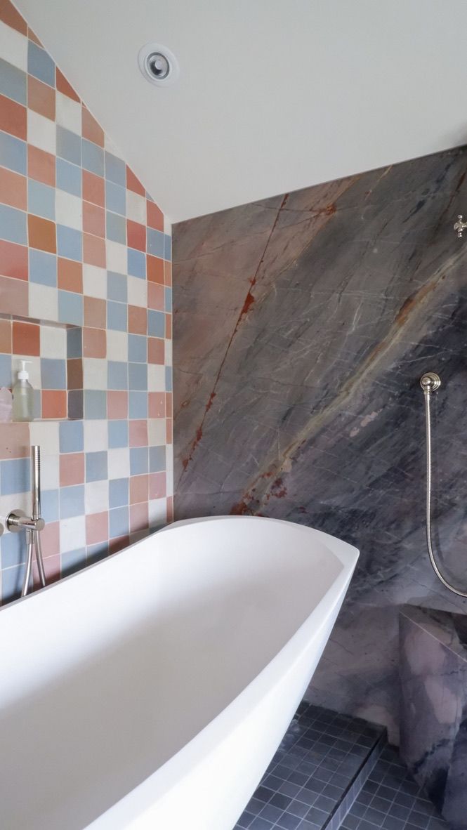 Freestanding white bathtub in a bathroom with tiled and marble walls; a shower head is present.