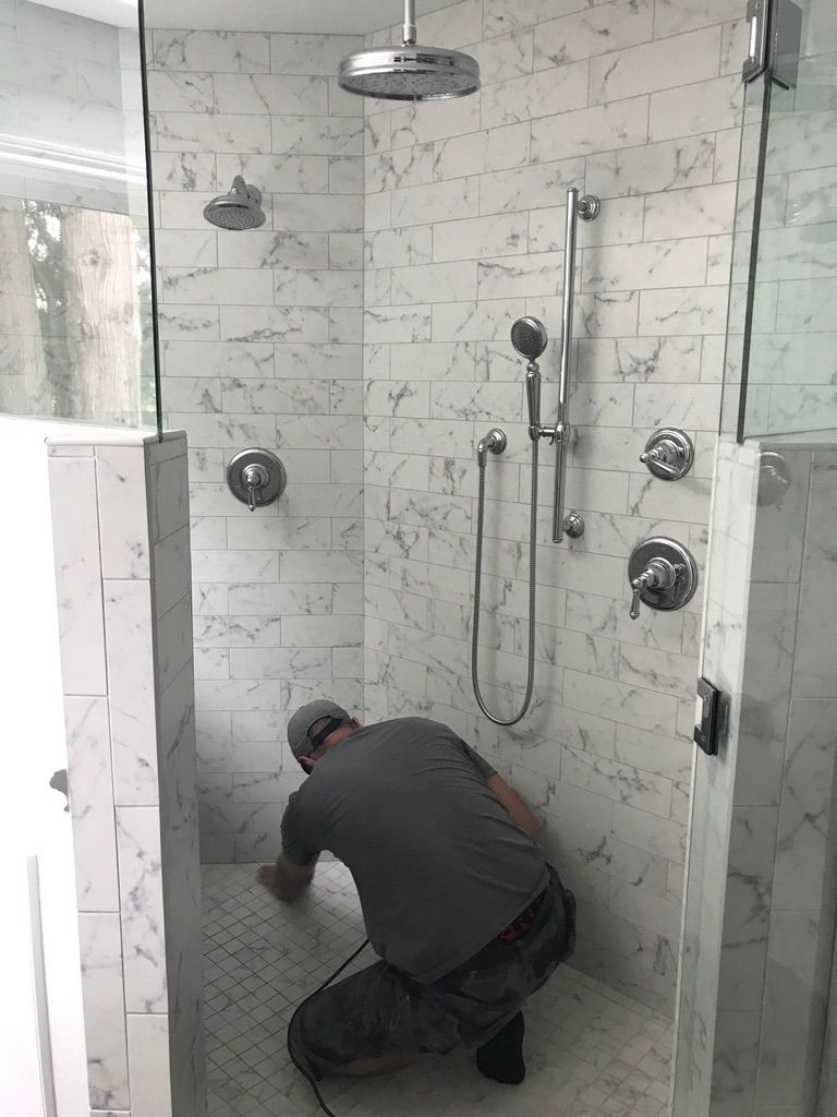 A person cleaning a white tiled shower with multiple shower heads, inside a bathroom.