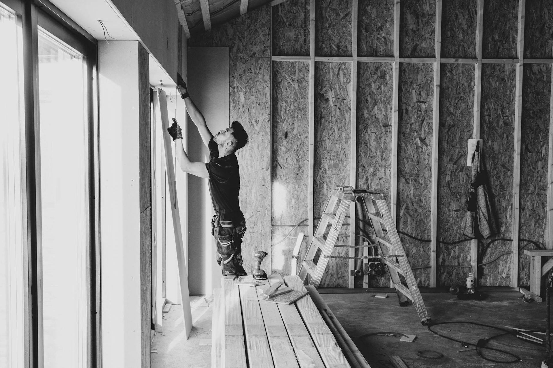 Construction worker inside a building, installing wall panels near a large window. Black and white photo.