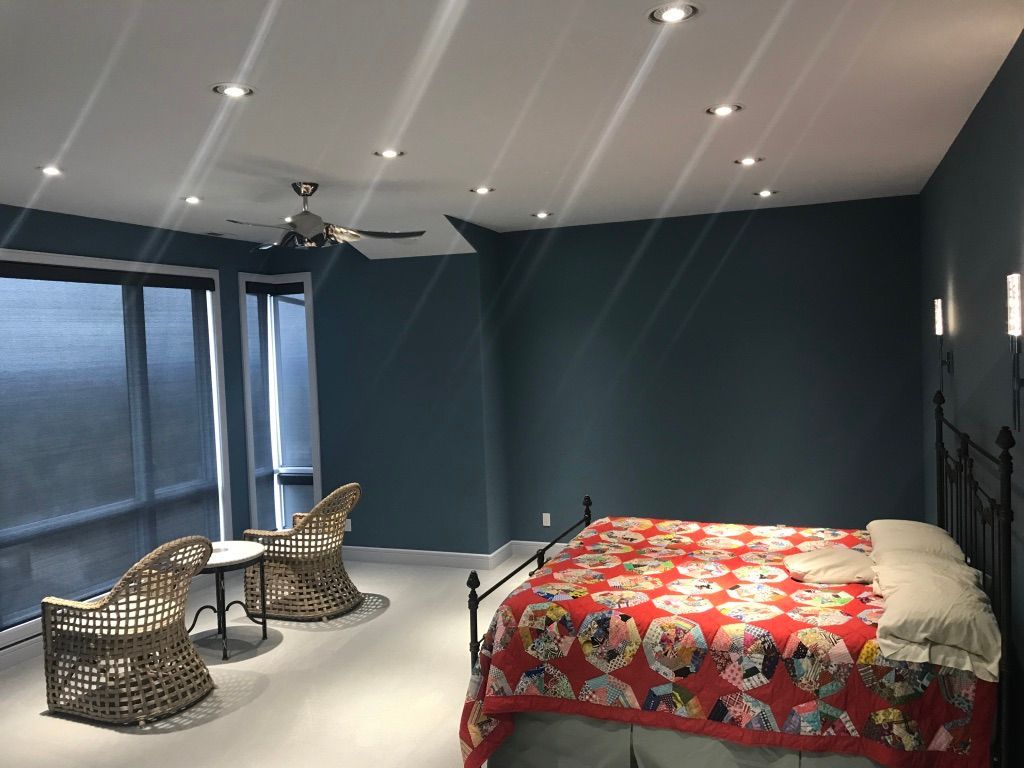 Bedroom with blue walls, a bed with a red quilt, wicker chairs, and a large window with blinds.