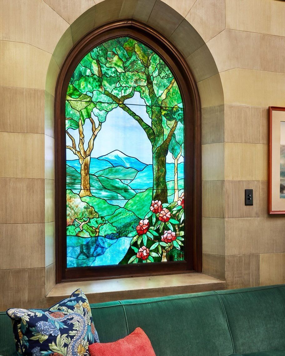Stained glass window with trees and a lake view, set in a stone wall. Green couch and pillows below.