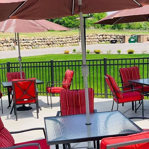 Outdoor patio with red chairs, tables, and brown umbrellas. Black fence, green lawn, and stone wall in background.