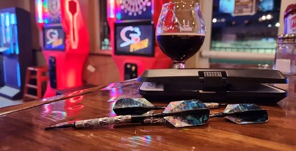 Dartboard darts and a beer glass on a bar, with arcade games in the background.