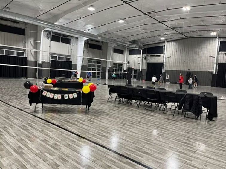 Birthday party setup in a gymnasium with balloons, cake, tables, chairs, and volleyball net.