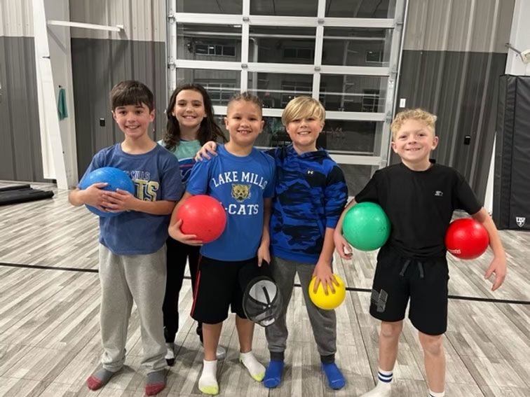 Five children hold colorful balls in a gymnasium, smiling at the camera.