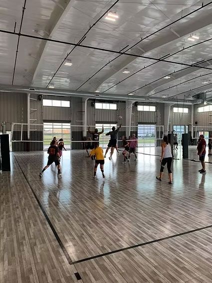 People playing volleyball in a large indoor gym with light wood floors and high ceilings.