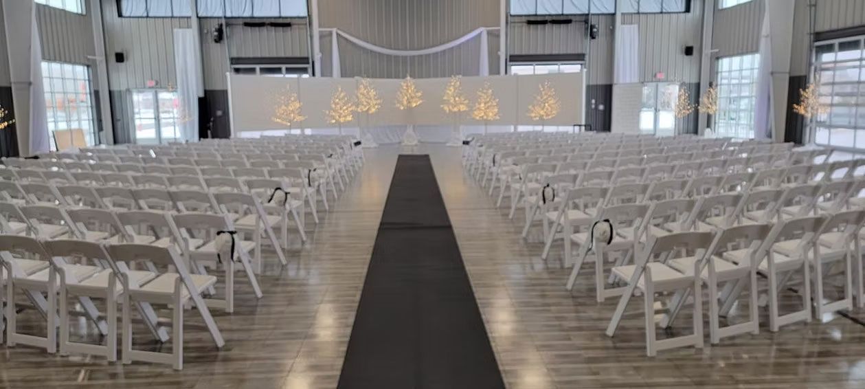 An indoor event space set up for a wedding, with rows of white chairs facing a decorated stage.