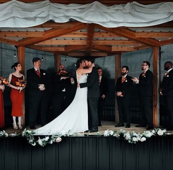Wedding ceremony: Bride and groom kissing under a wooden structure; wedding party looks on.