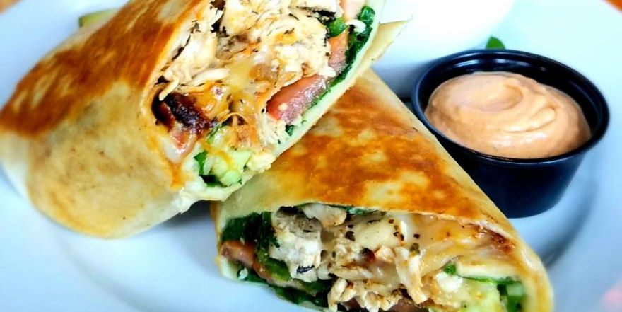 A grilled chicken wrap cut in half on a white plate, served with a side of creamy orange dipping sauce.