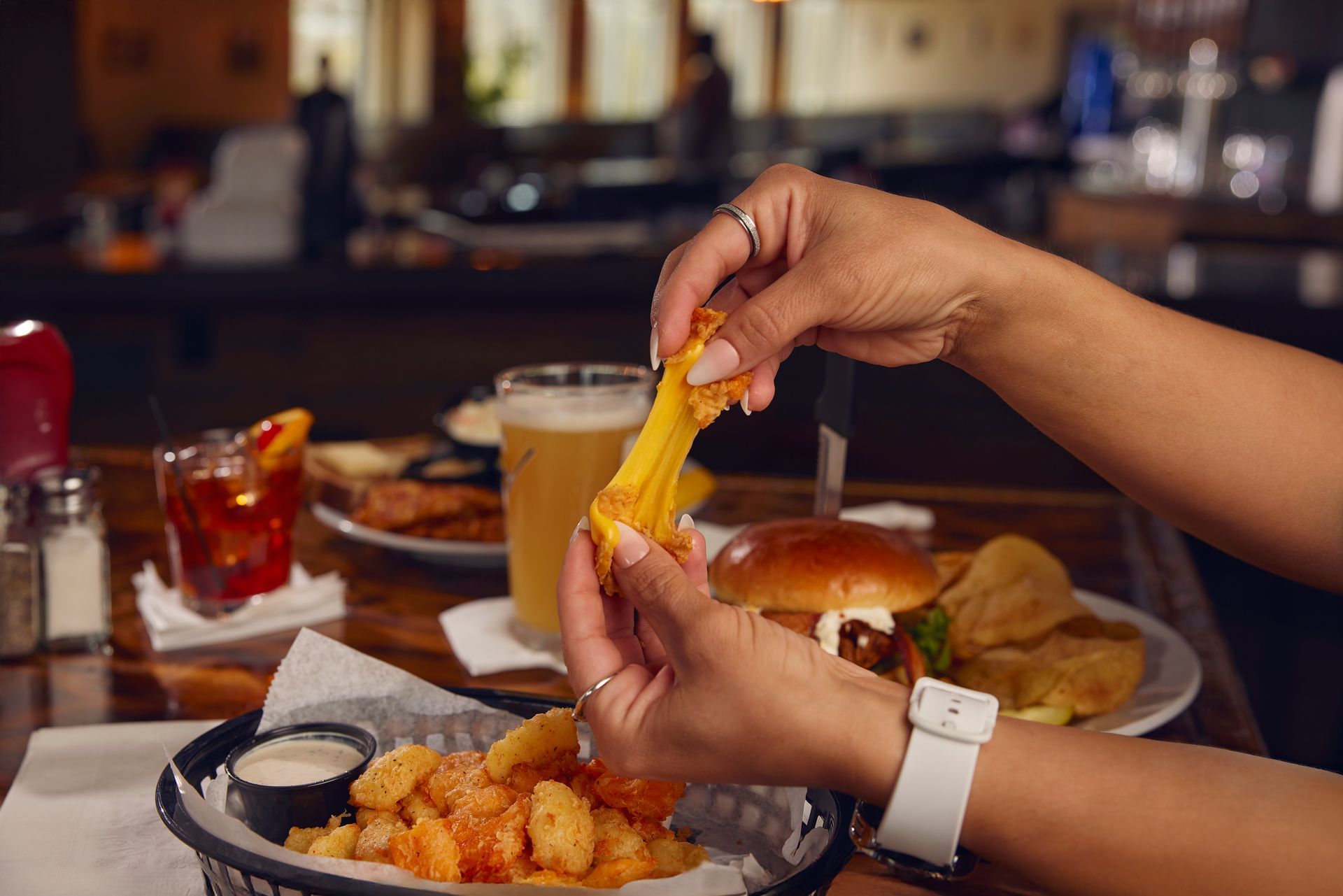 Hands tearing a fried snack over a table with burgers, shrimp, and drinks in a restaurant