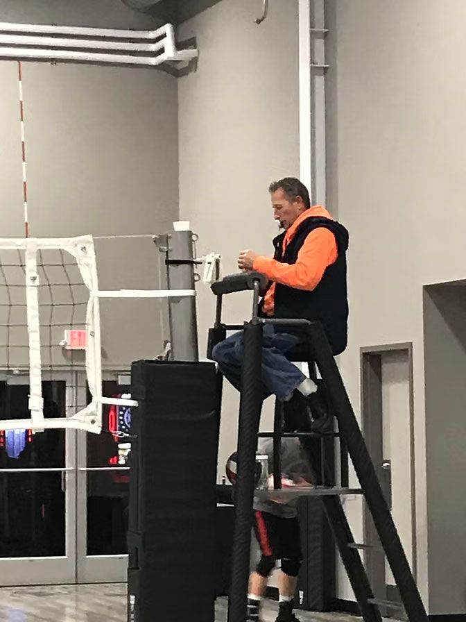 Man adjusting volleyball net, sitting in referee stand. Indoor gym setting, wearing a vest and jeans.