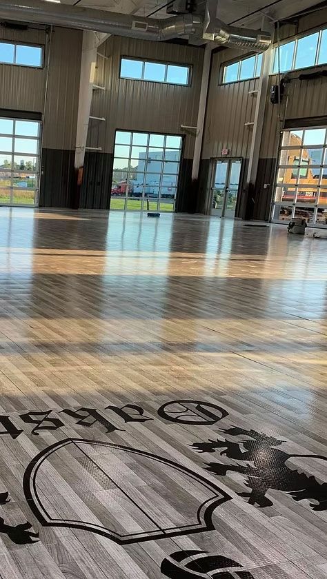 Wooden floor inside a large building with high ceilings and windows, sunlight casting shadows.
