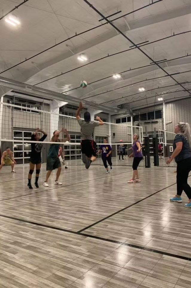 Volleyball game: Player jumping to hit the ball, other players ready to play on a wooden court.