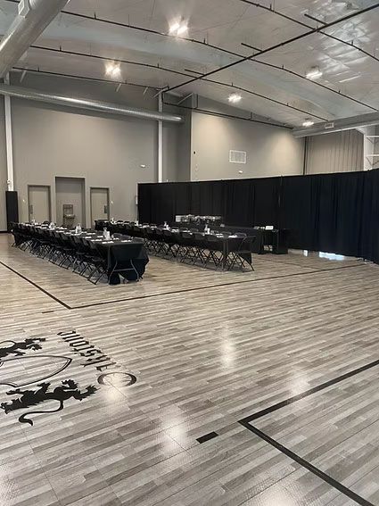 Tables set up in a large indoor space. Black table cloths, black curtains, light wood-look floor, basketball court lines.