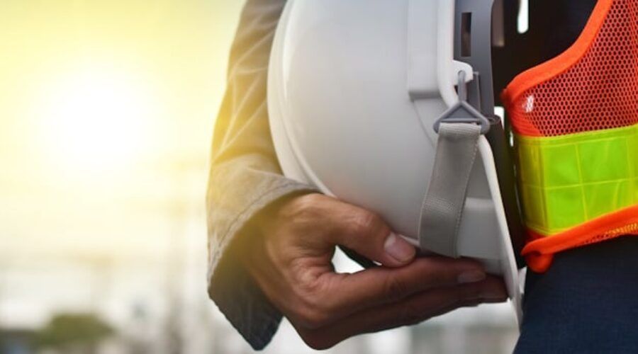 Construction worker holding a hard hat, wearing a safety vest with sunlight in the background.