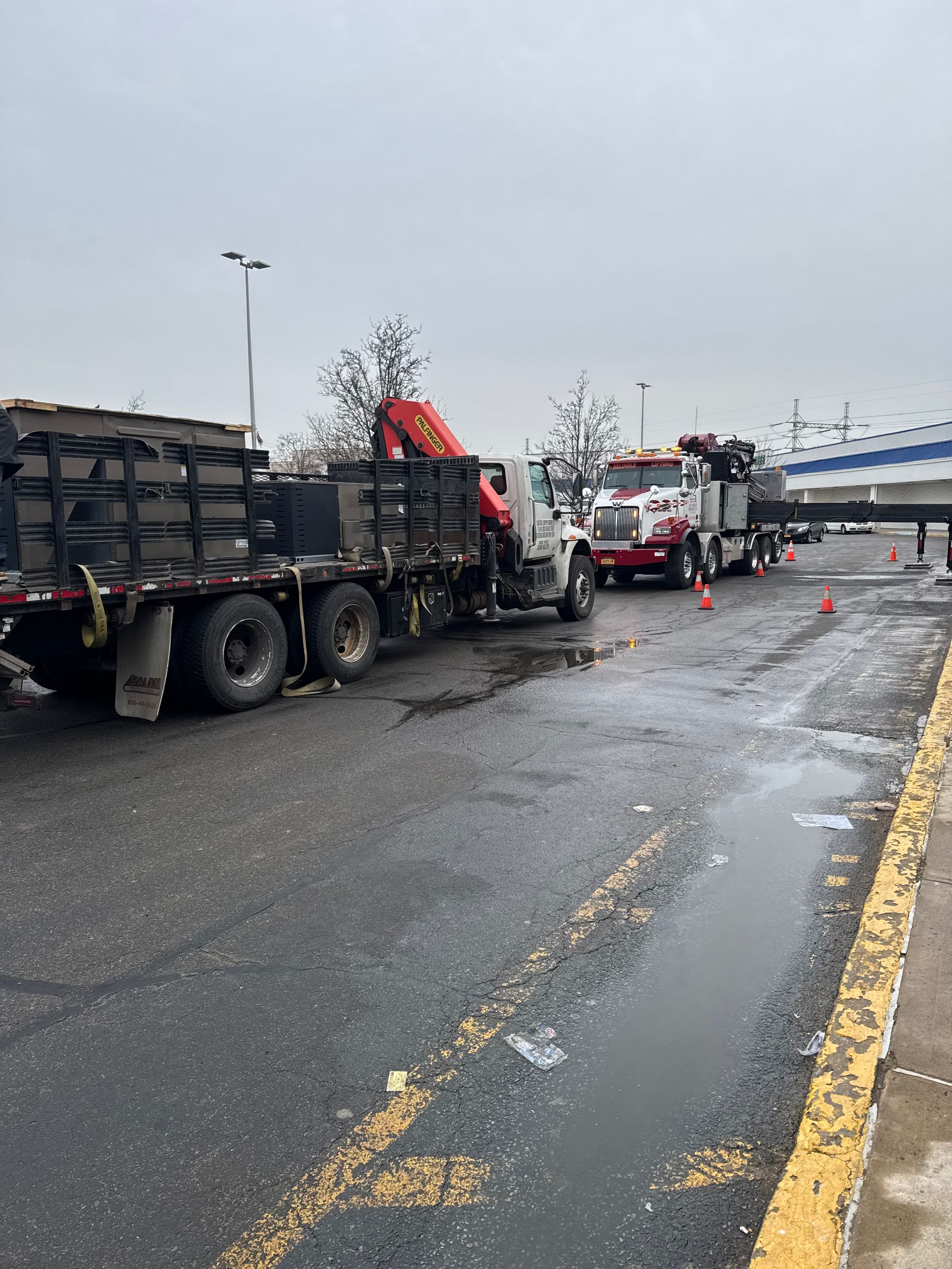 Truck loading equipment onto a flatbed trailer in a wet parking lot.