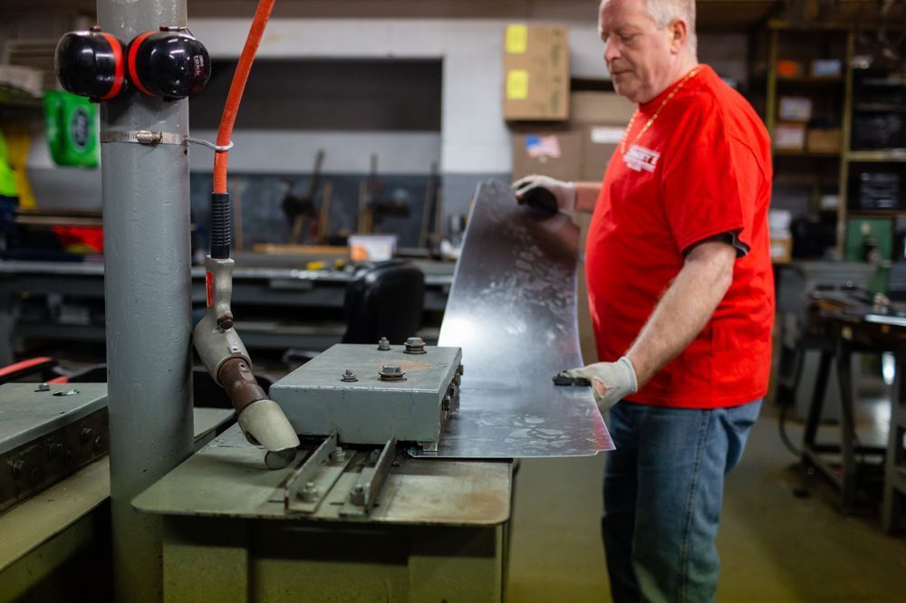 Man in red shirt working with a metal sheet and industrial machinery in a workshop.