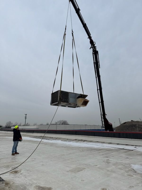 A crane lifting an HVAC unit; a worker in a hard hat watches. Overcast sky.