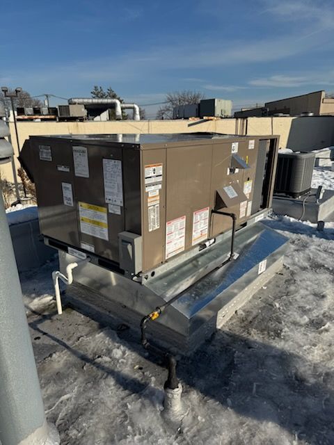 Rooftop HVAC unit on a building, brown with labels, surrounded by snow and pipes against a blue sky.