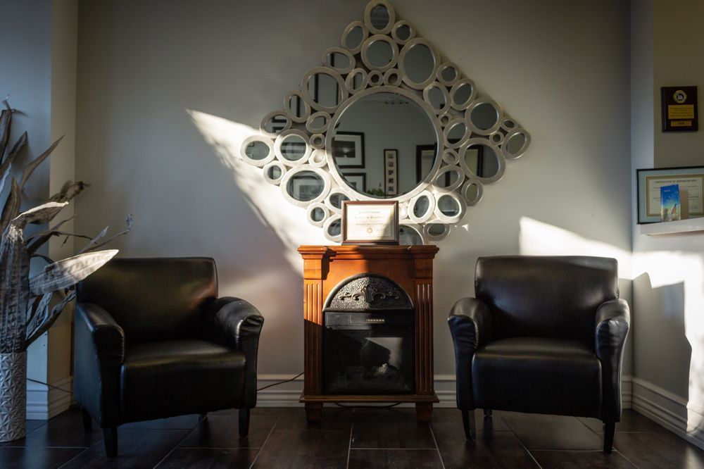 Two black leather armchairs face a fireplace beneath a large, decorative mirror; bright sunlight streams in.