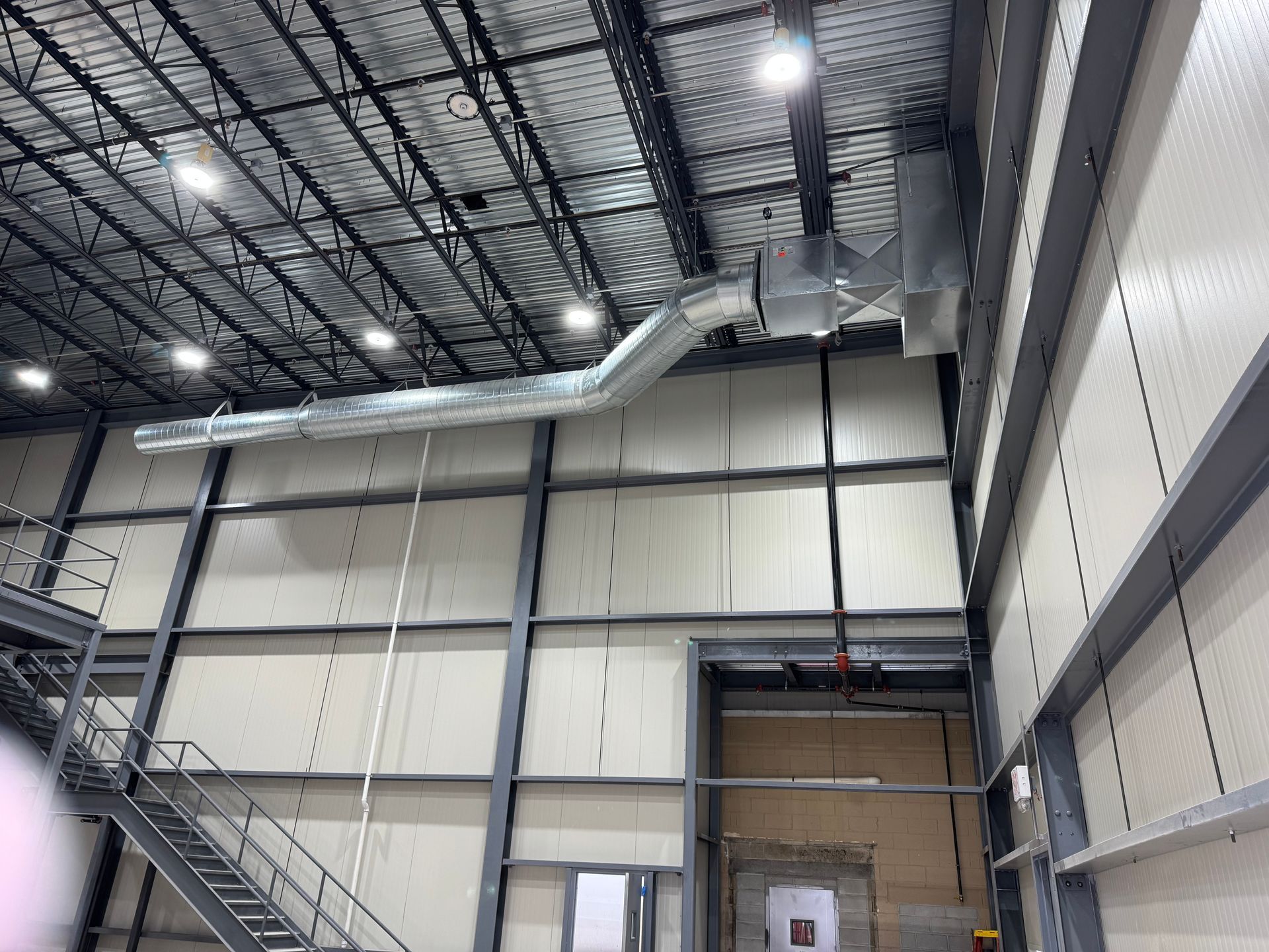 Interior of a large industrial building with metal beams, ductwork, and a staircase. White walls and ceiling with overhead lighting.