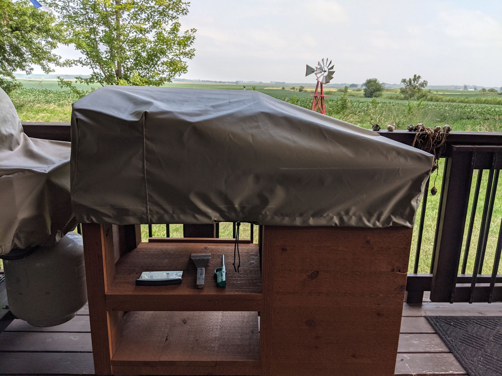 A grill with a cover on it sits on a deck with a windmill in the background