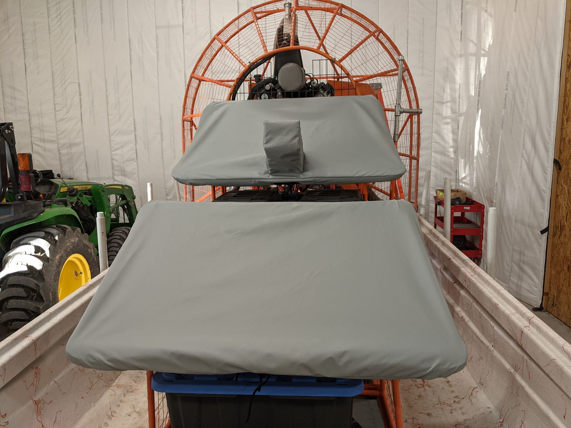 A boat with a cover on it is sitting in a garage next to a john deere tractor.