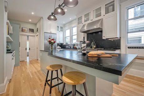 Modern kitchen with island, black countertop, light wood floor, stools, white cabinets, and stainless appliances.