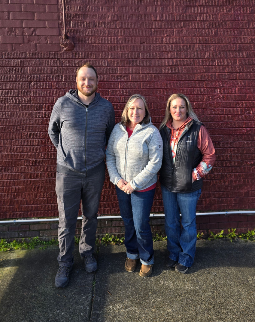 Three people standing in front of a textured, reddish-brown brick wall on a sunny day.