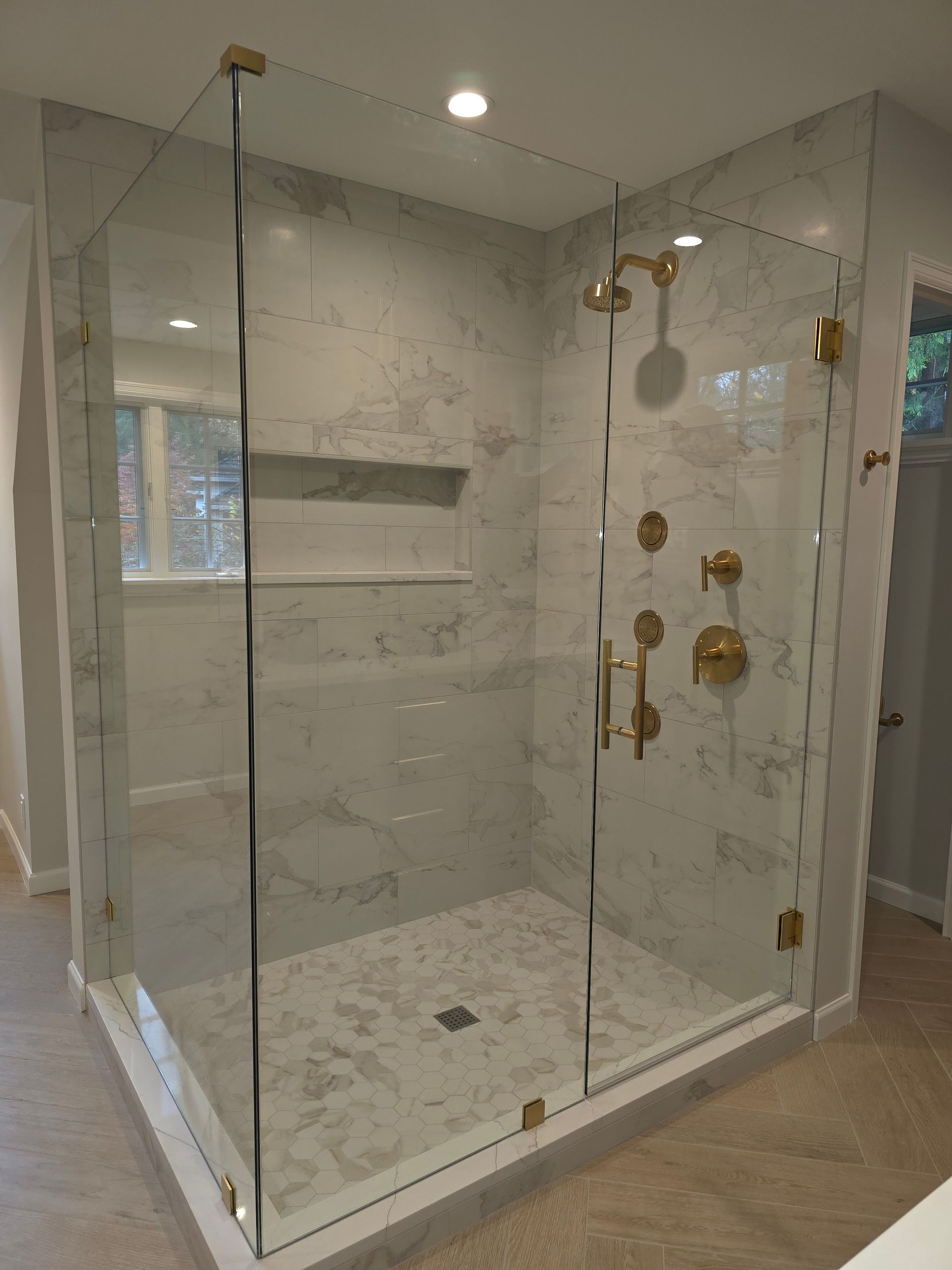 A walk-in glass shower with marble-tiled walls and floor, featuring gold hardware and a built-in recessed shelf.